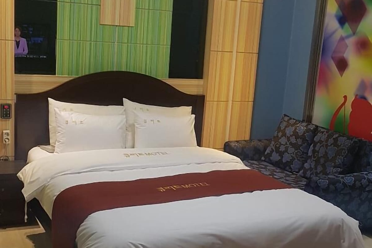 A hotel room with a large bed, a wooden headboard, a green wall panel, and a colorful abstract painting.