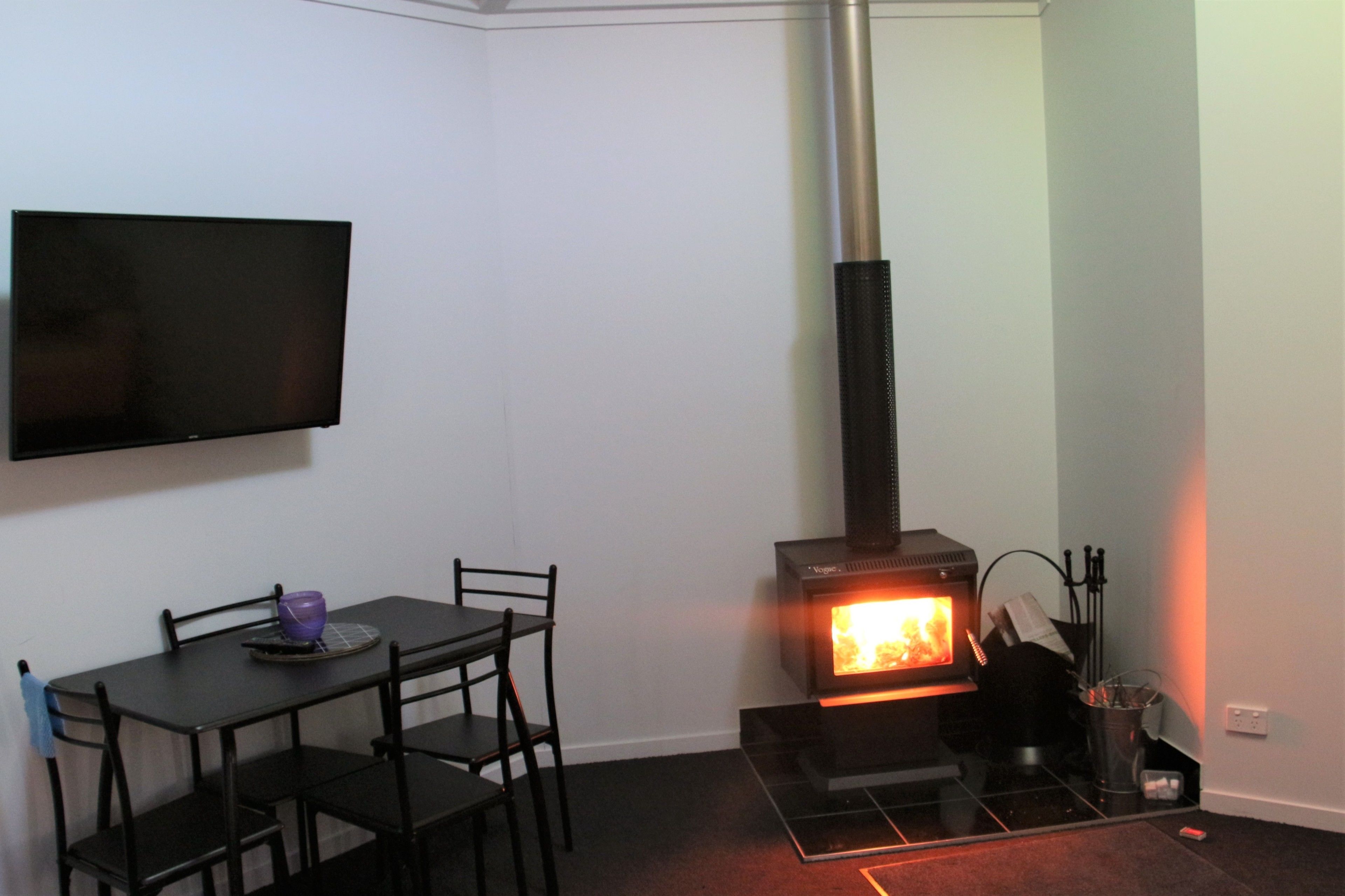 A room with a black table and chairs, a flat-screen TV mounted on the wall, and a wood-burning stove.