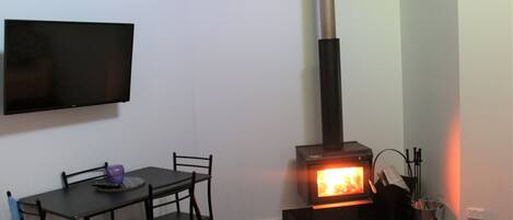 A room with a black table and chairs, a flat-screen TV mounted on the wall, and a wood-burning stove.