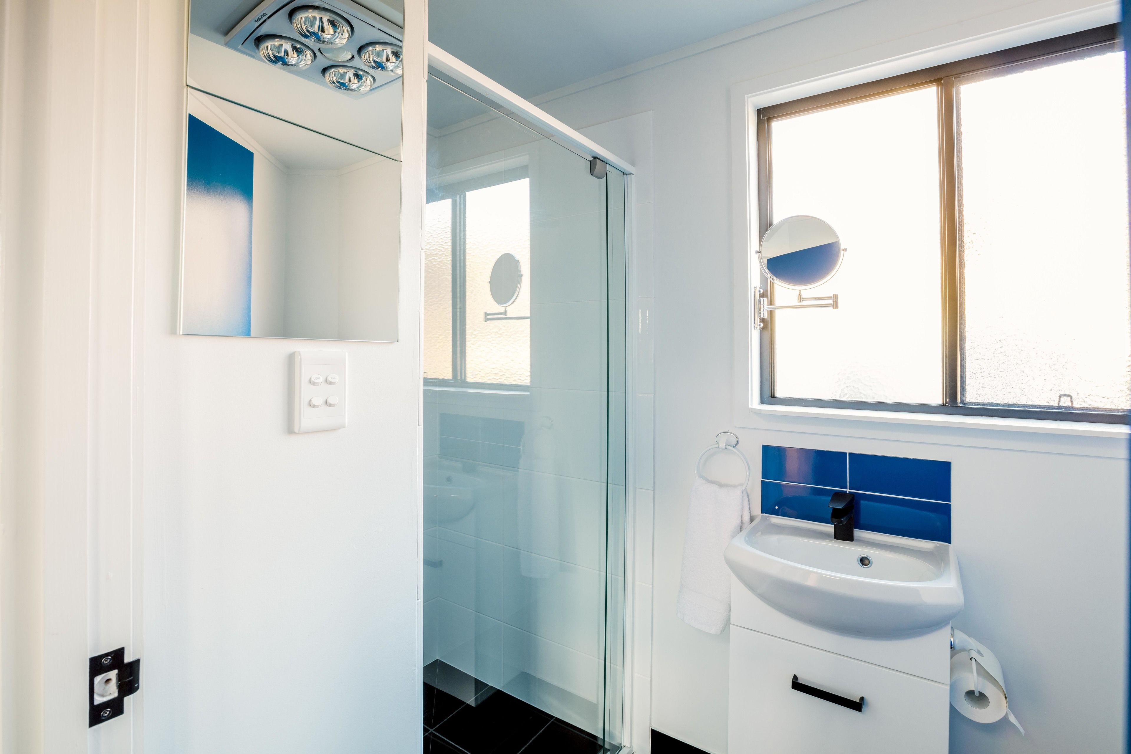 Un baño moderno con cabina de ducha de vidrio, un lavamanos blanco y un salpicadero azul, y una ventana sobre el lavamanos.