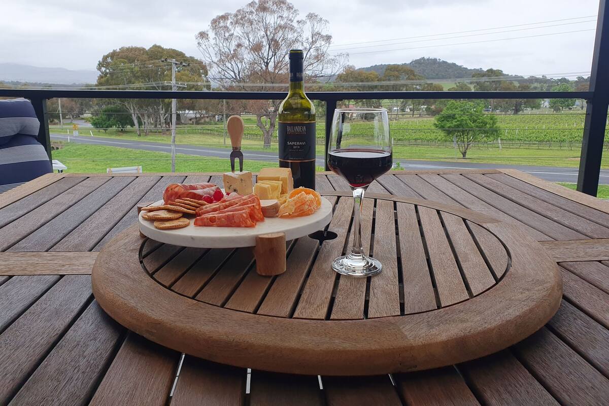 A wooden table with a cheese board, crackers, and a bottle of wine on a balcony with a view of a road and trees.