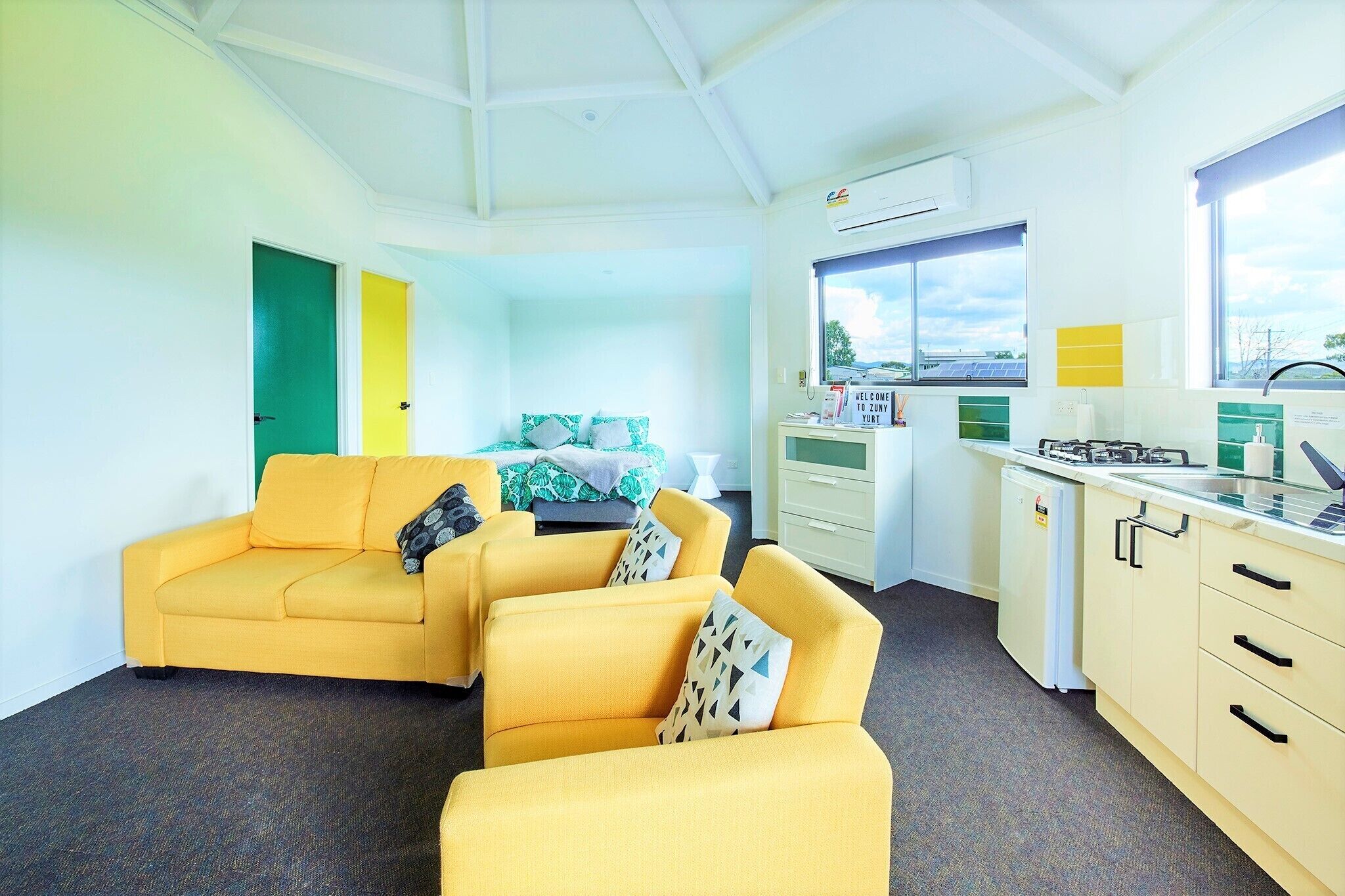 A bright room with yellow sofas, a bed with green and white bedding, a small kitchenette, and a window with a view.