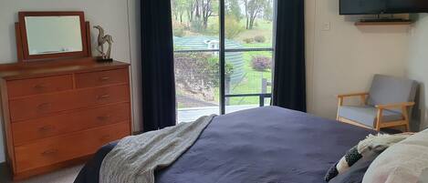 A bedroom with a bed, a dresser, a chair, a television, and a balcony with a view of greenery.