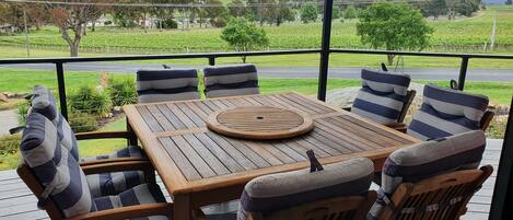 A covered outdoor seating area with a wooden table and striped cushioned chairs, overlooking a vineyard and a road.