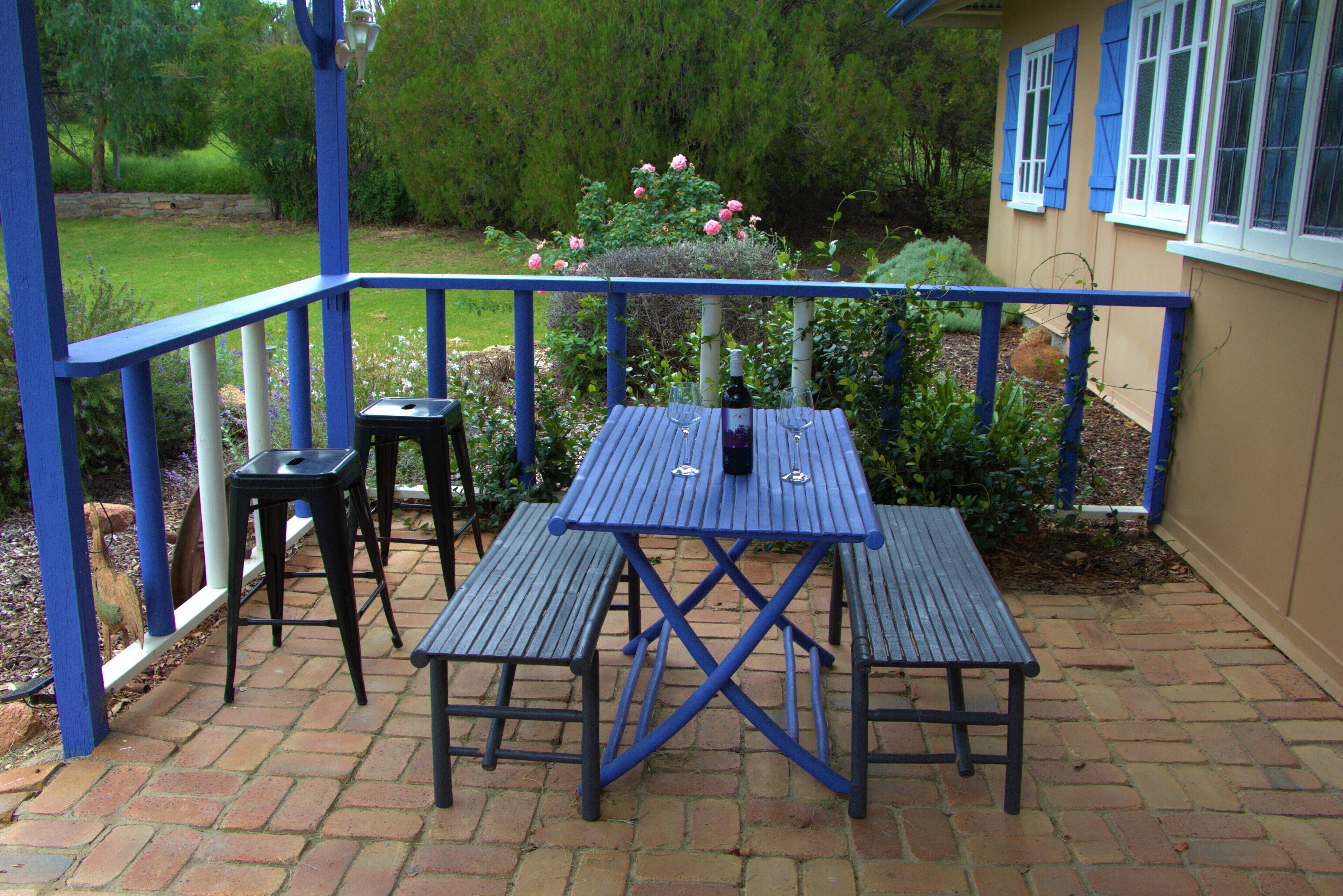 Eine Terrasse mit blauem Tisch und Stühlen, umgeben von einem Backsteinboden und einem Garten mit rosafarbenen Blumen.