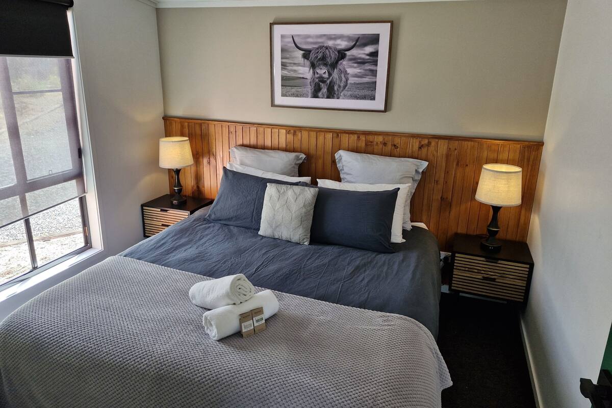 A neatly made bed with a grey and white duvet, two pillows, and a rolled white towel. There are two bedside tables with lamps, a framed picture on the wall, and a window with blinds.