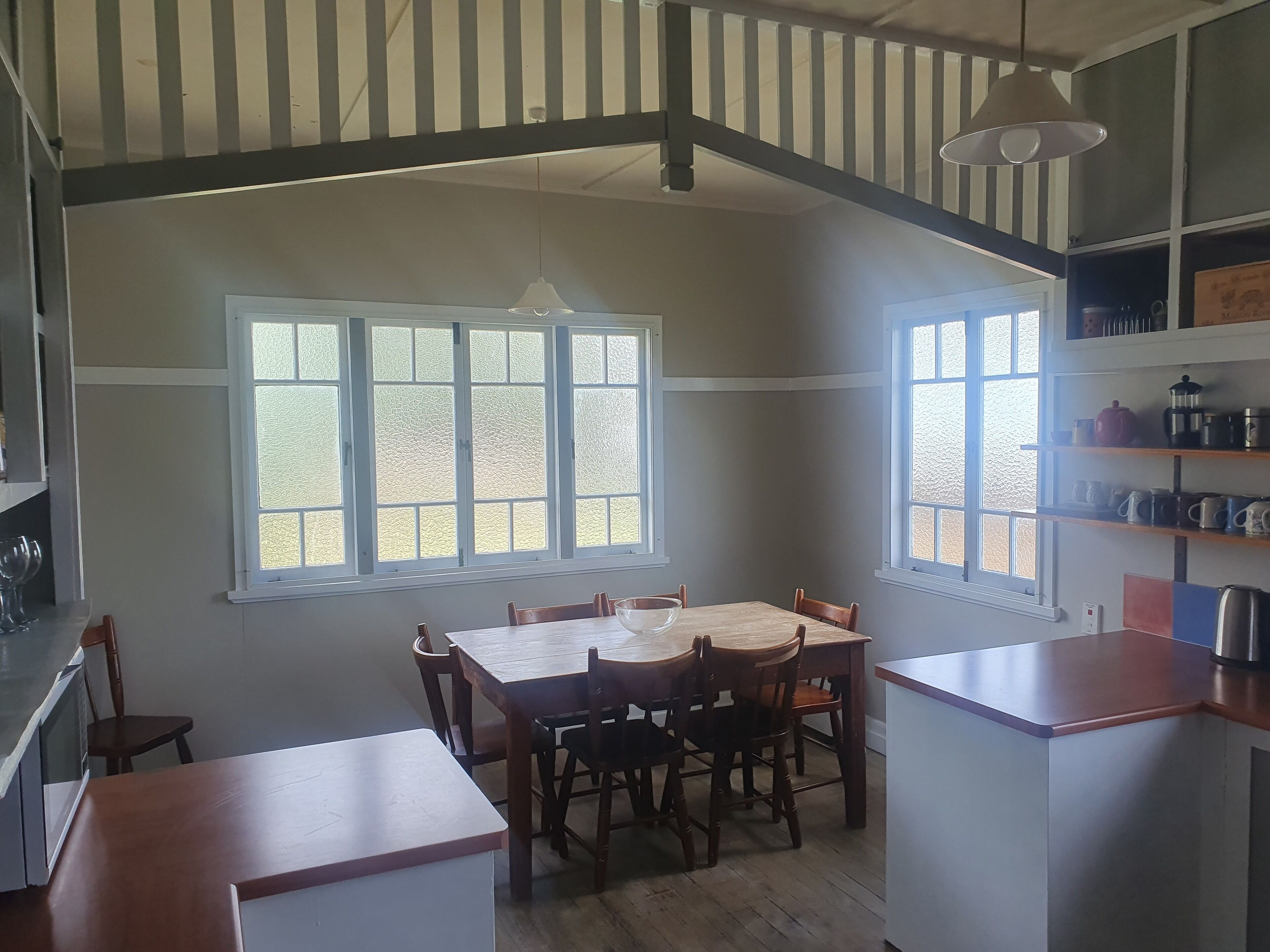 A dining area with a wooden table and chairs, large windows, and a kitchen area with shelves.