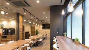 Un café moderno con largas mesas de madera y sillas blancas, una máquina de café y un amplio ventanal con persianas.
