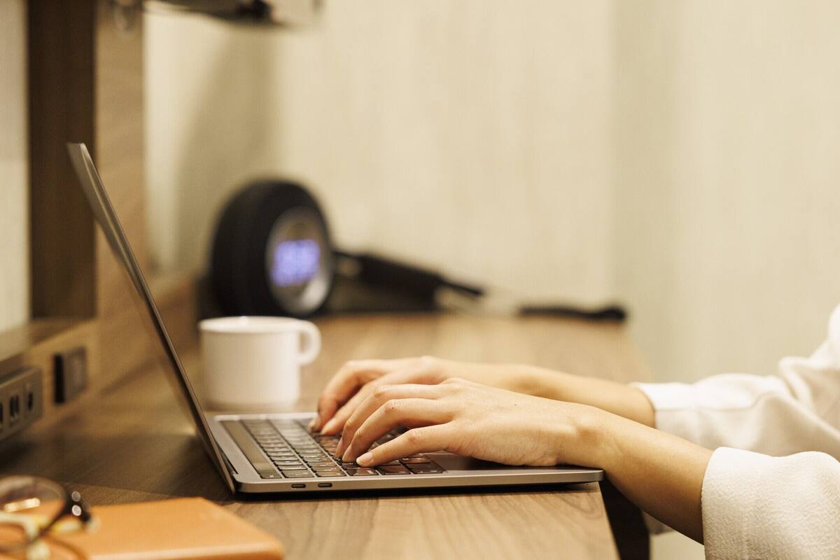 A person using a laptop on a wooden desk with a white mug and glasses nearby.