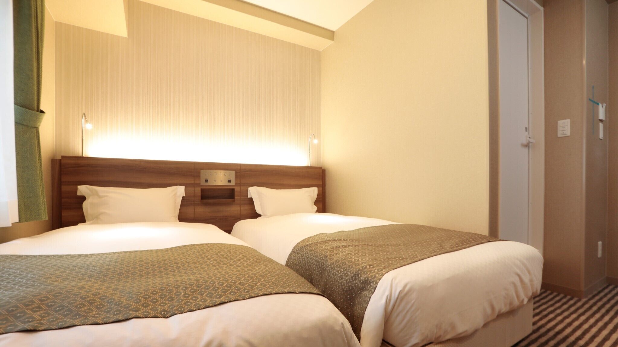 A hotel room with two beds, a wooden headboard, a wall-mounted light, and a door.