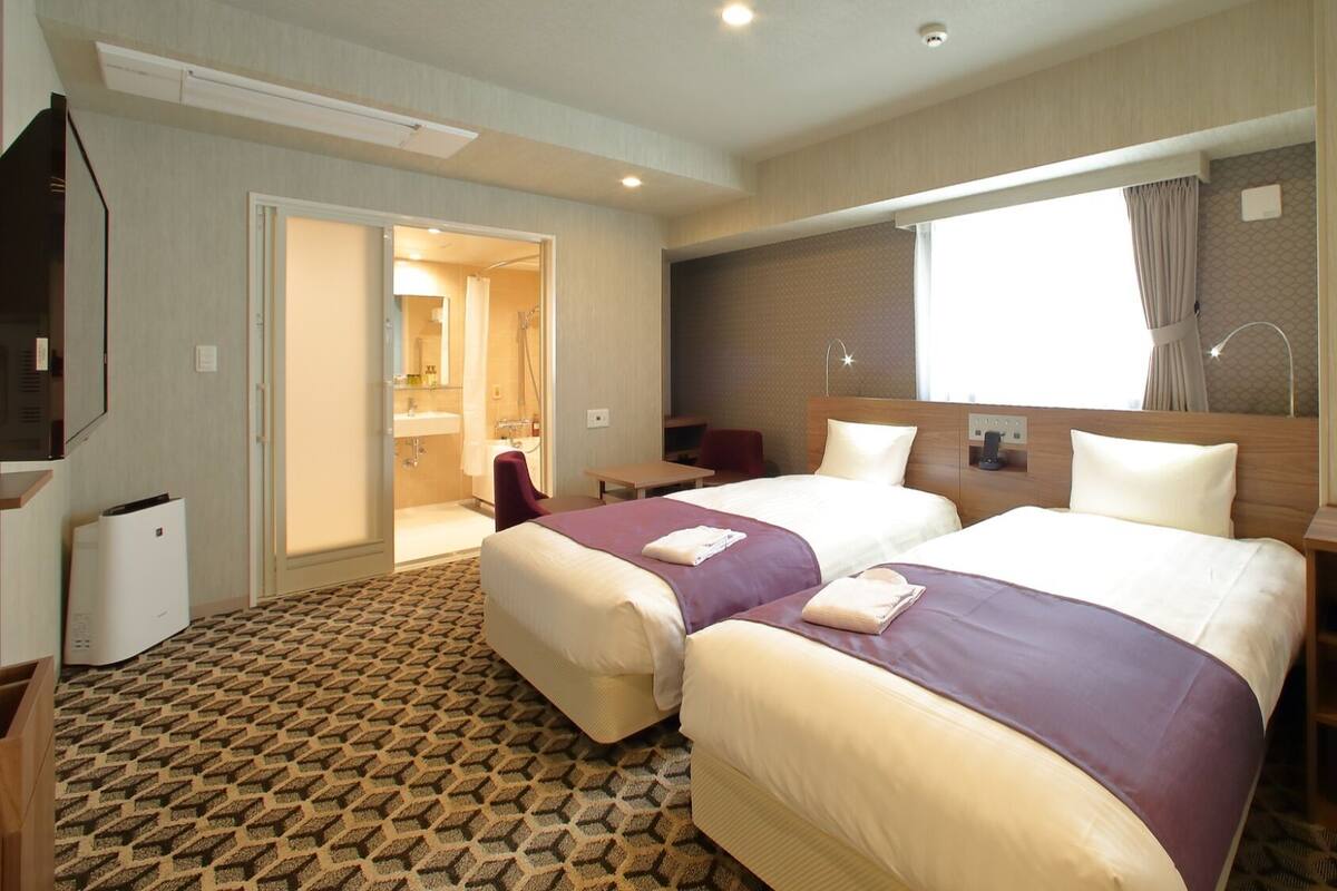 A hotel room with two beds, a desk, a chair, a TV, and a bathroom visible through an open door.