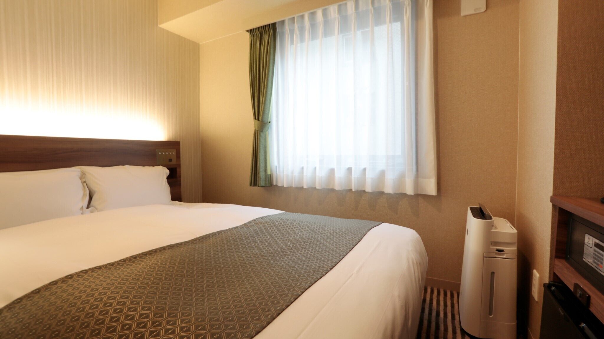 A hotel room with a large bed, a bedside heater, a window with sheer curtains, and a wall-mounted air conditioner.