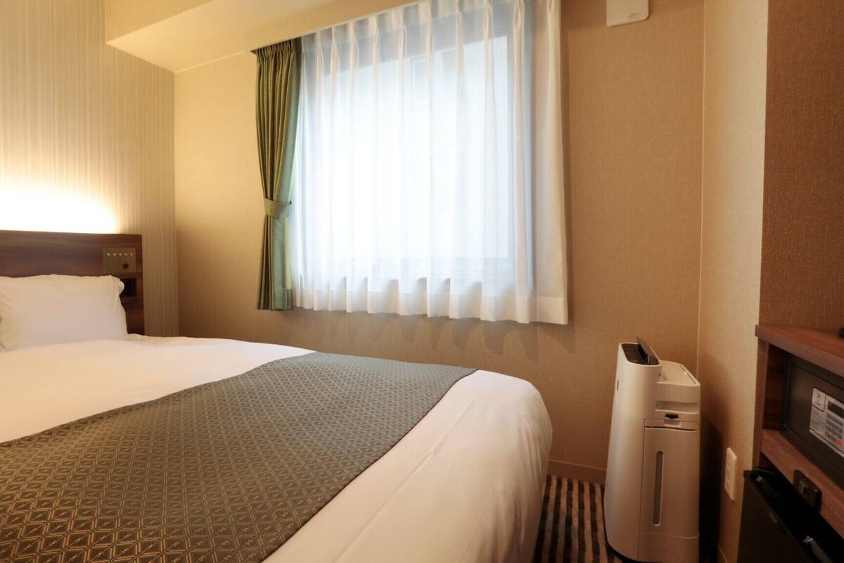 A hotel room with a large bed, a bedside heater, a window with sheer curtains, and a wall-mounted air conditioner.
