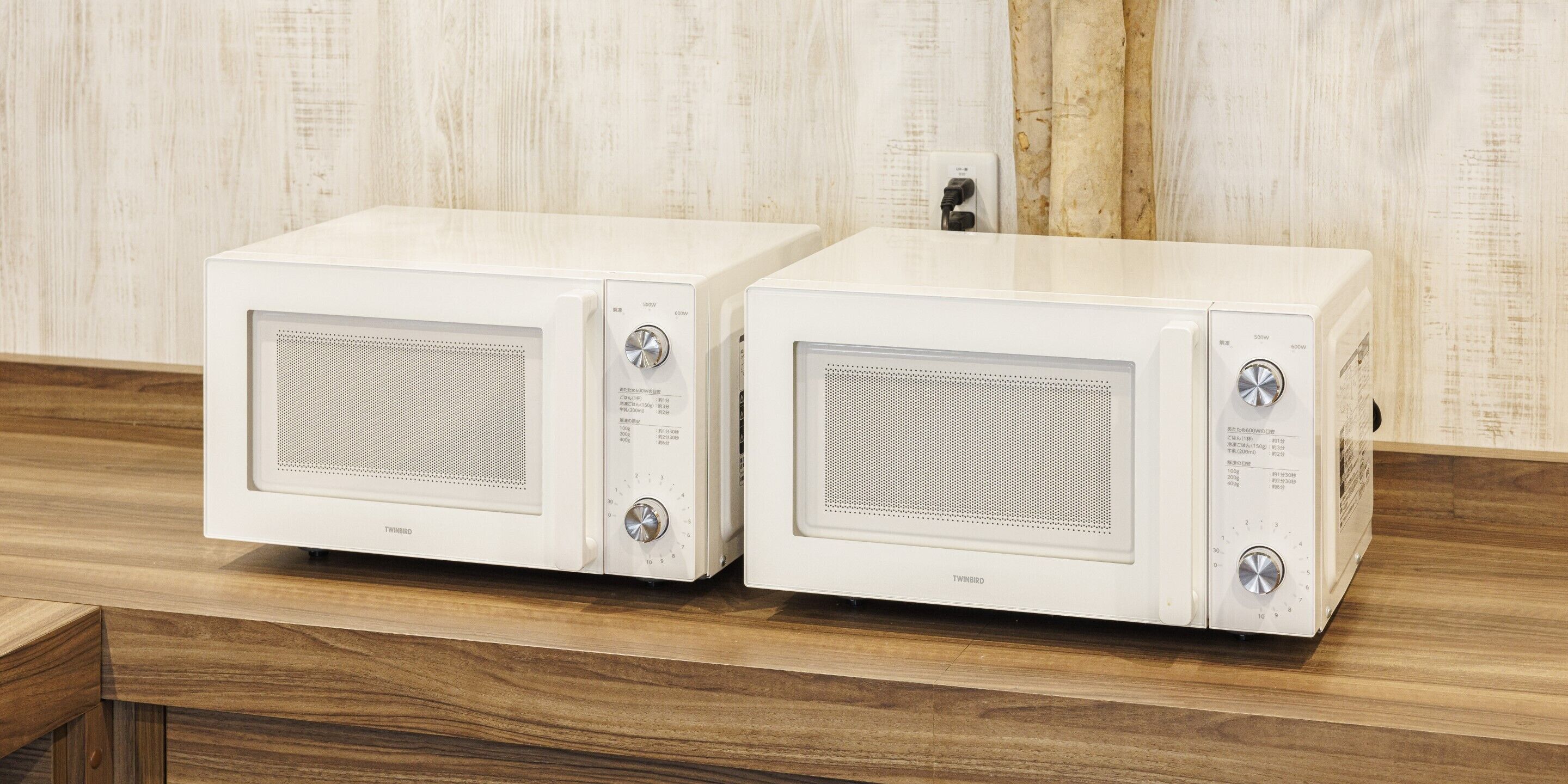 Deux fours à micro-ondes blancs de la marque TWIRBIRD sont posés sur un plan de travail en bois, avec un mur en bois clair en arrière-plan.