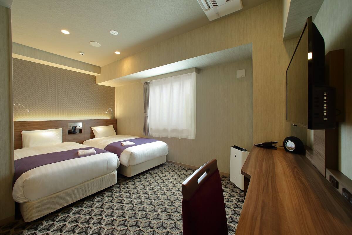 A hotel room with two beds, a desk, a TV, and a window with curtains.
