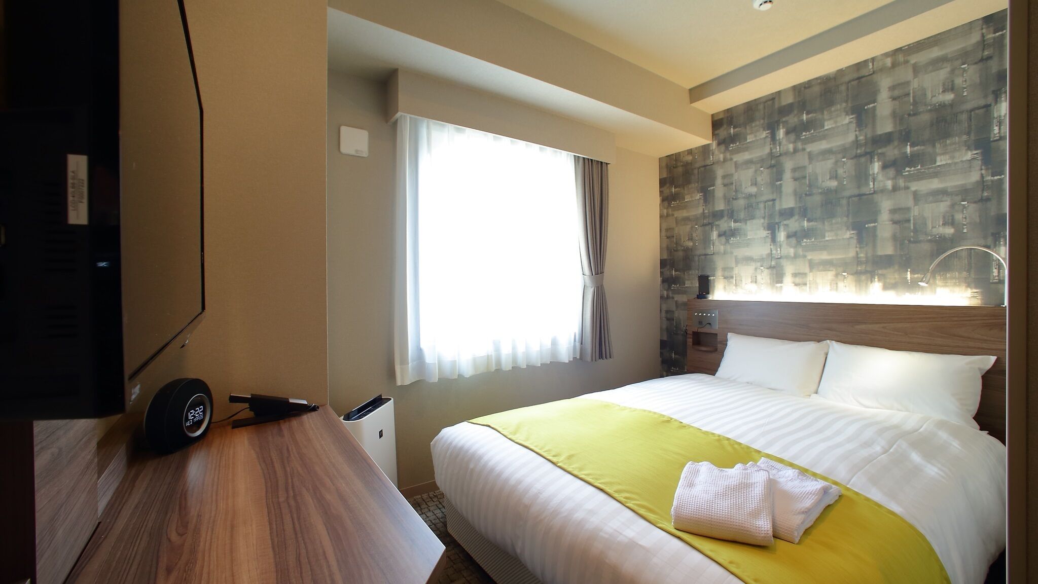 A hotel room with a bed, a desk, a TV, a window with curtains, and a wall with a textured pattern.