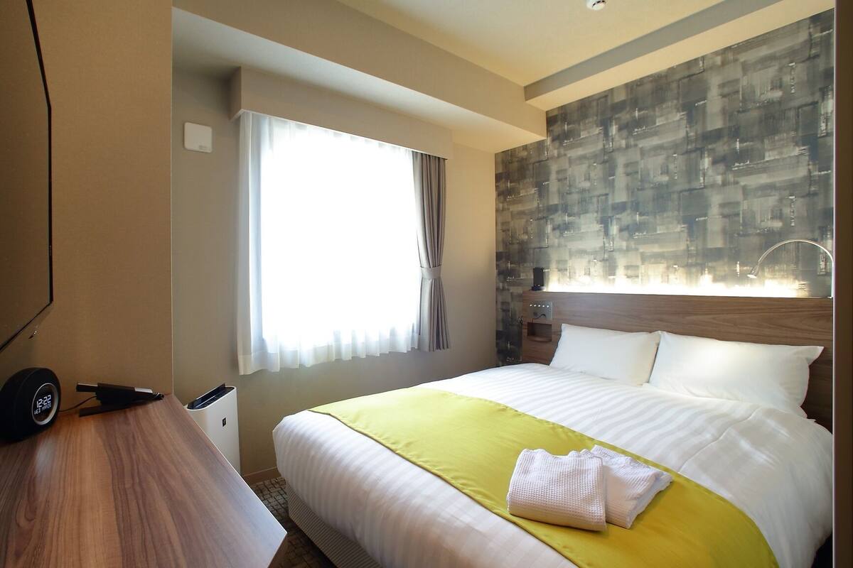 A hotel room with a bed, a desk, a TV, a window with curtains, and a wall with a textured pattern.