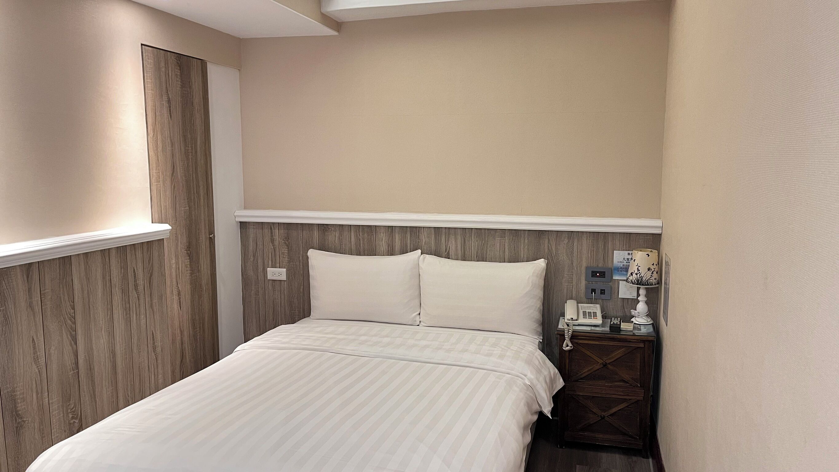 A neatly made bed with a wooden headboard, a bedside table with a lamp, and a wall-mounted phone.