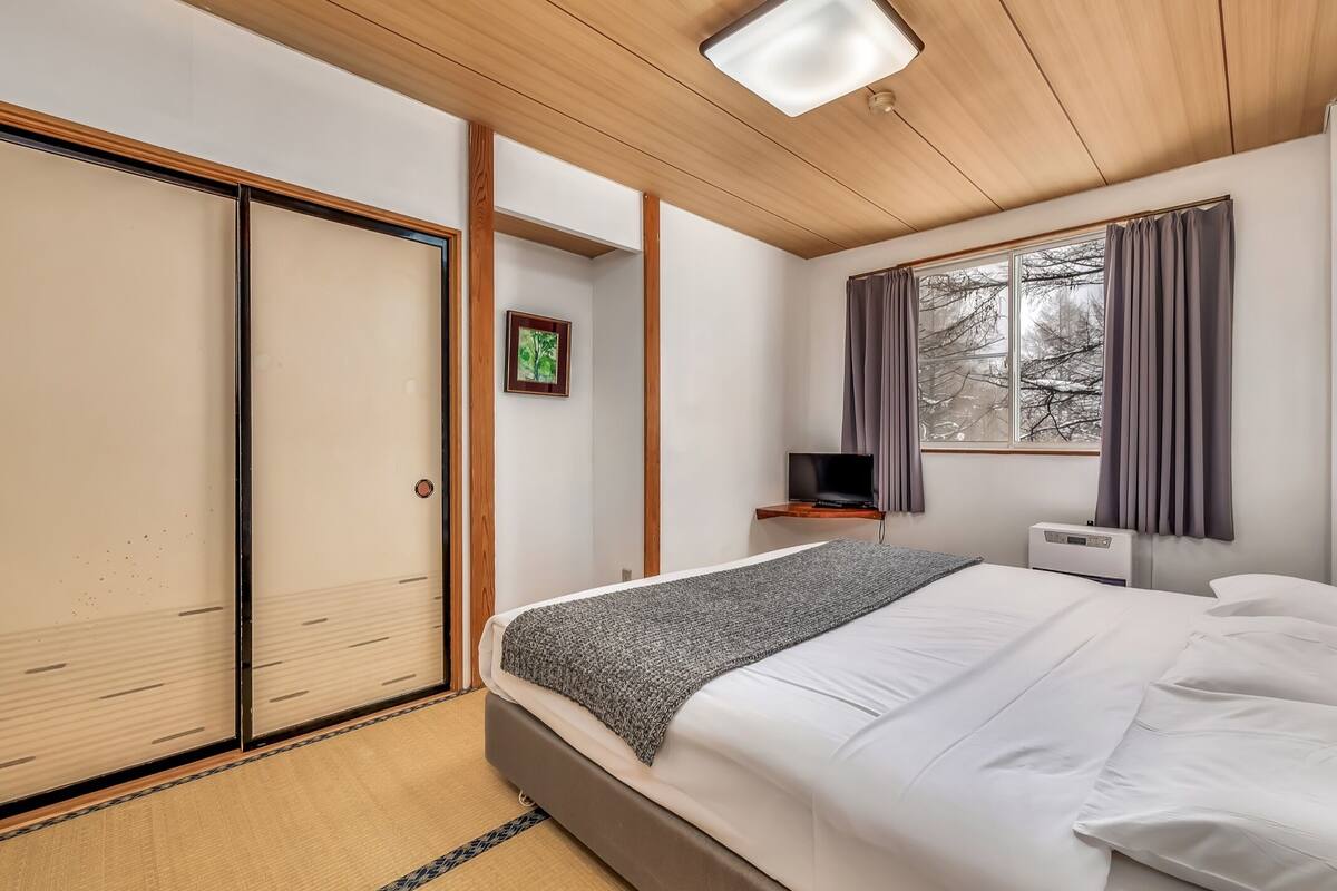A bedroom with a bed, a TV on a shelf, a heater, and a window with curtains.