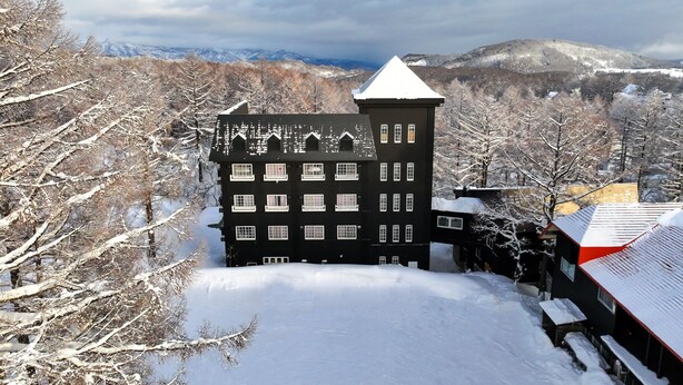 Un bâtiment noir doté de nombreuses fenêtres et d’une tour, entouré d’arbres et de montagnes enneigés en arrière-plan.