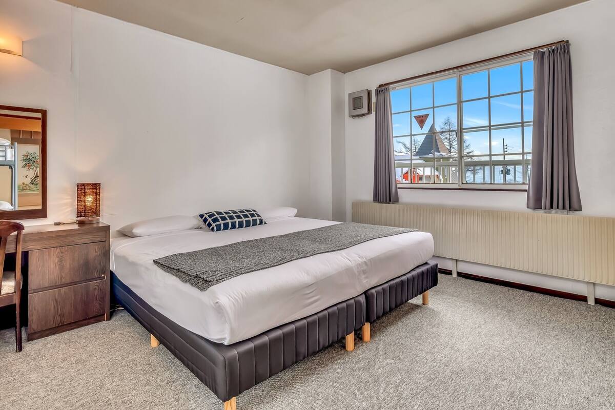A bedroom with a bed, a bedside table, a lamp, a window with curtains, and a radiator.