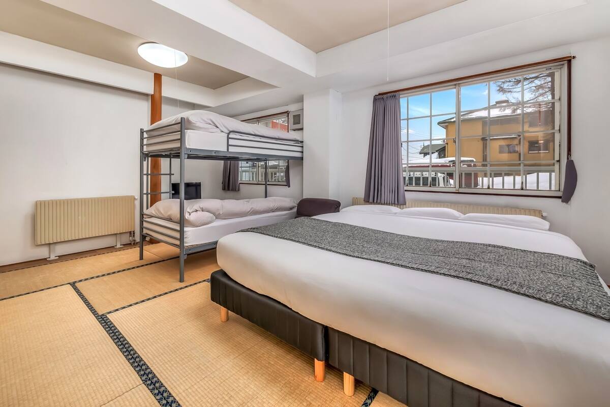 A modern hotel room with a bunk bed, a large bed, a window with a view of the outside, and a ceiling light.