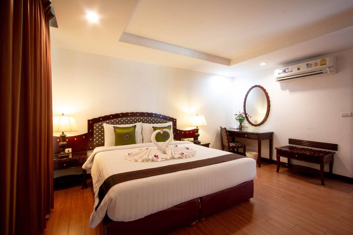 A hotel room with a large bed, two bedside tables with lamps, a wooden bench, a desk with a chair, and an air conditioning unit on the wall.