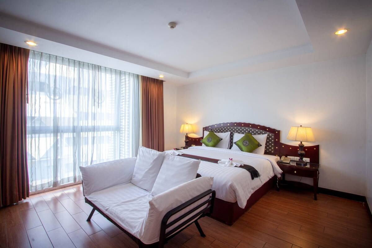 A hotel room with a bed, two bedside tables with lamps, a sofa, and a large window with curtains.