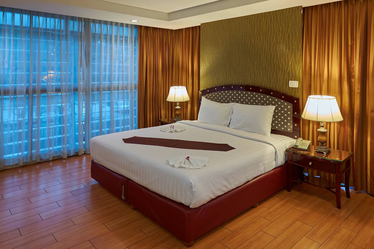 A hotel room with a large bed, two bedside tables with lamps, a wooden floor, and a view through sheer curtains.