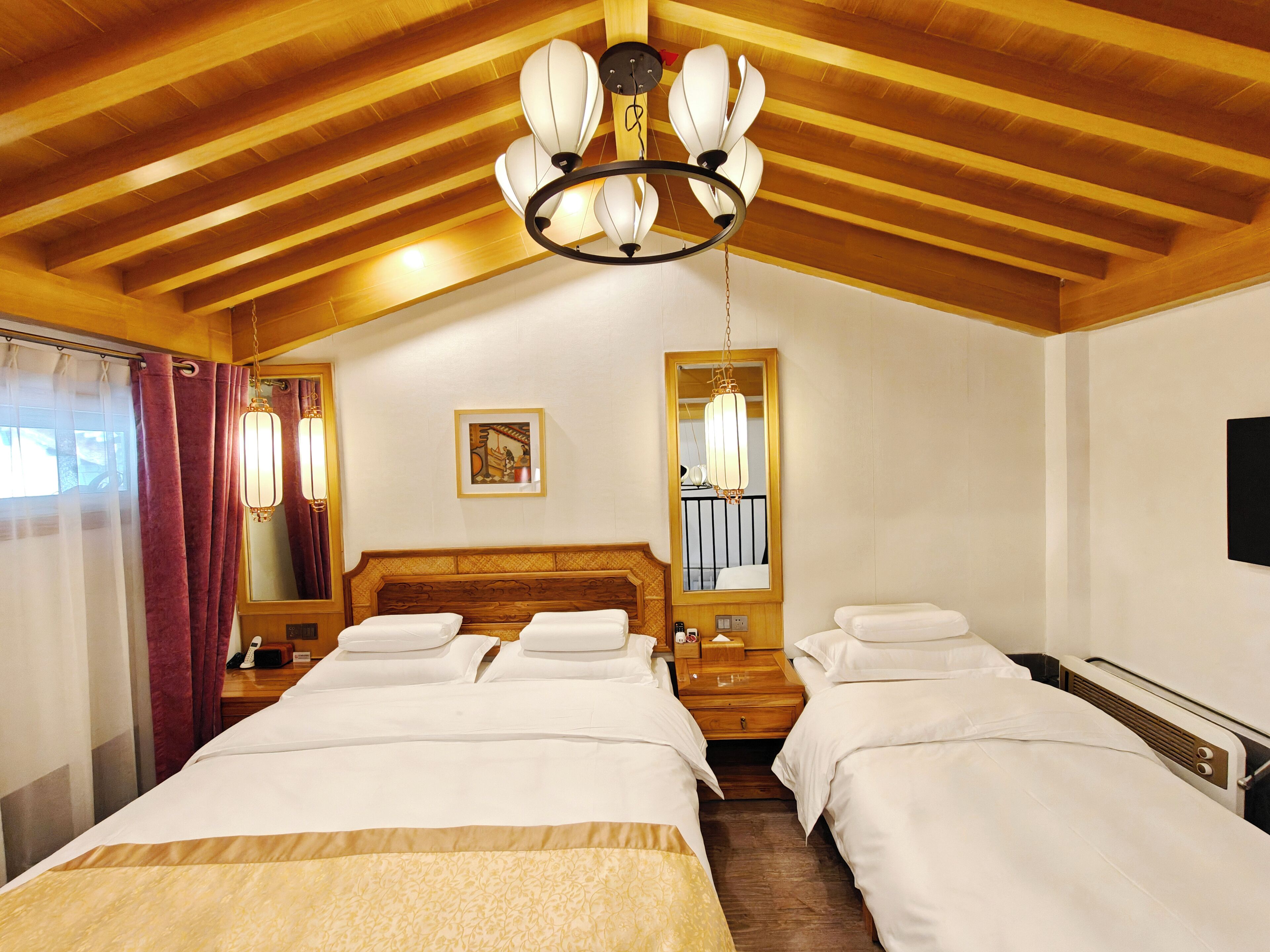 A hotel room with two beds, wooden ceiling beams, a chandelier, a large mirror, and a window with curtains.