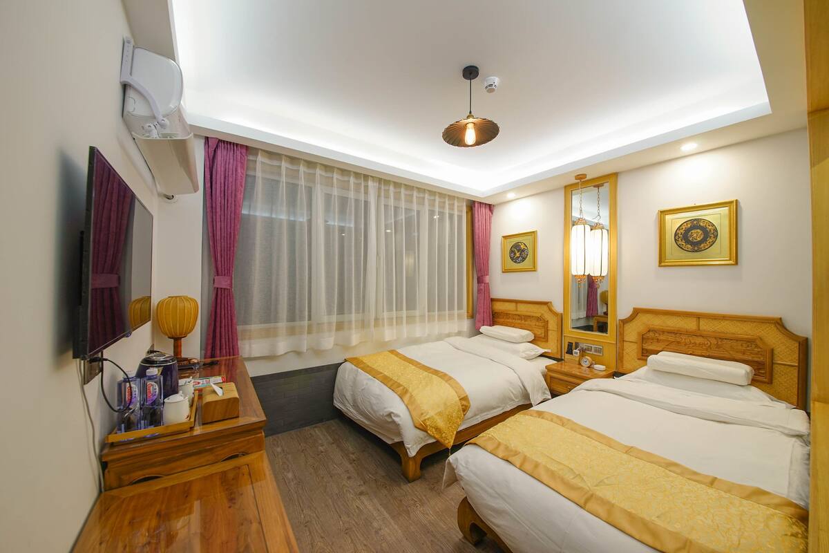 Habitación de hotel con dos camas, un tocador de madera, un escritorio con lámpara, un ventanal con cortinas traslúcidas y una lámpara de techo.