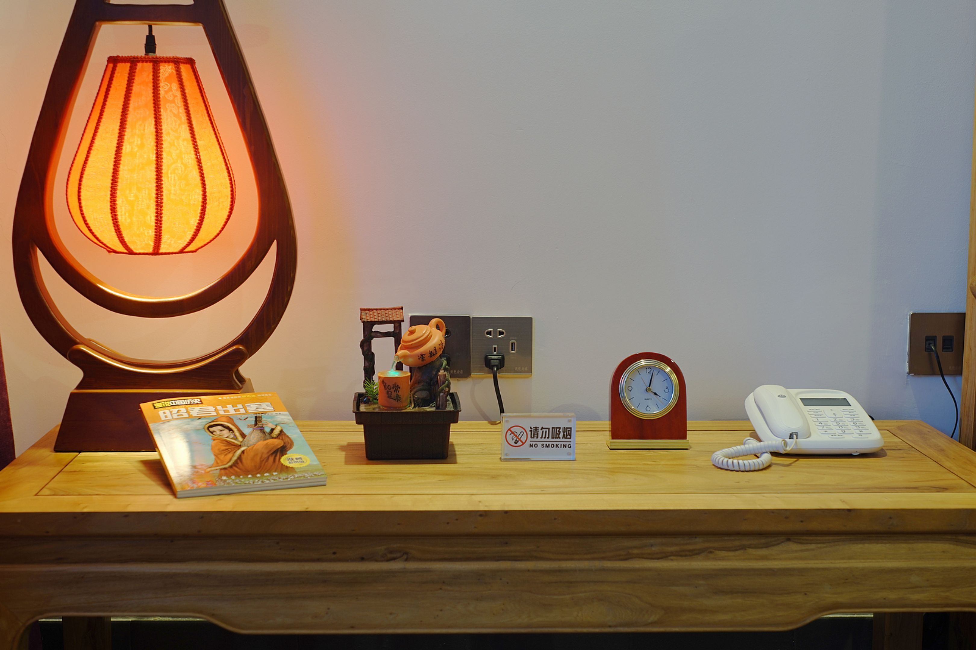 Un escritorio de madera con una lámpara, un libro, una figurita, un reloj, un teléfono y un cartel de prohibido fumar.