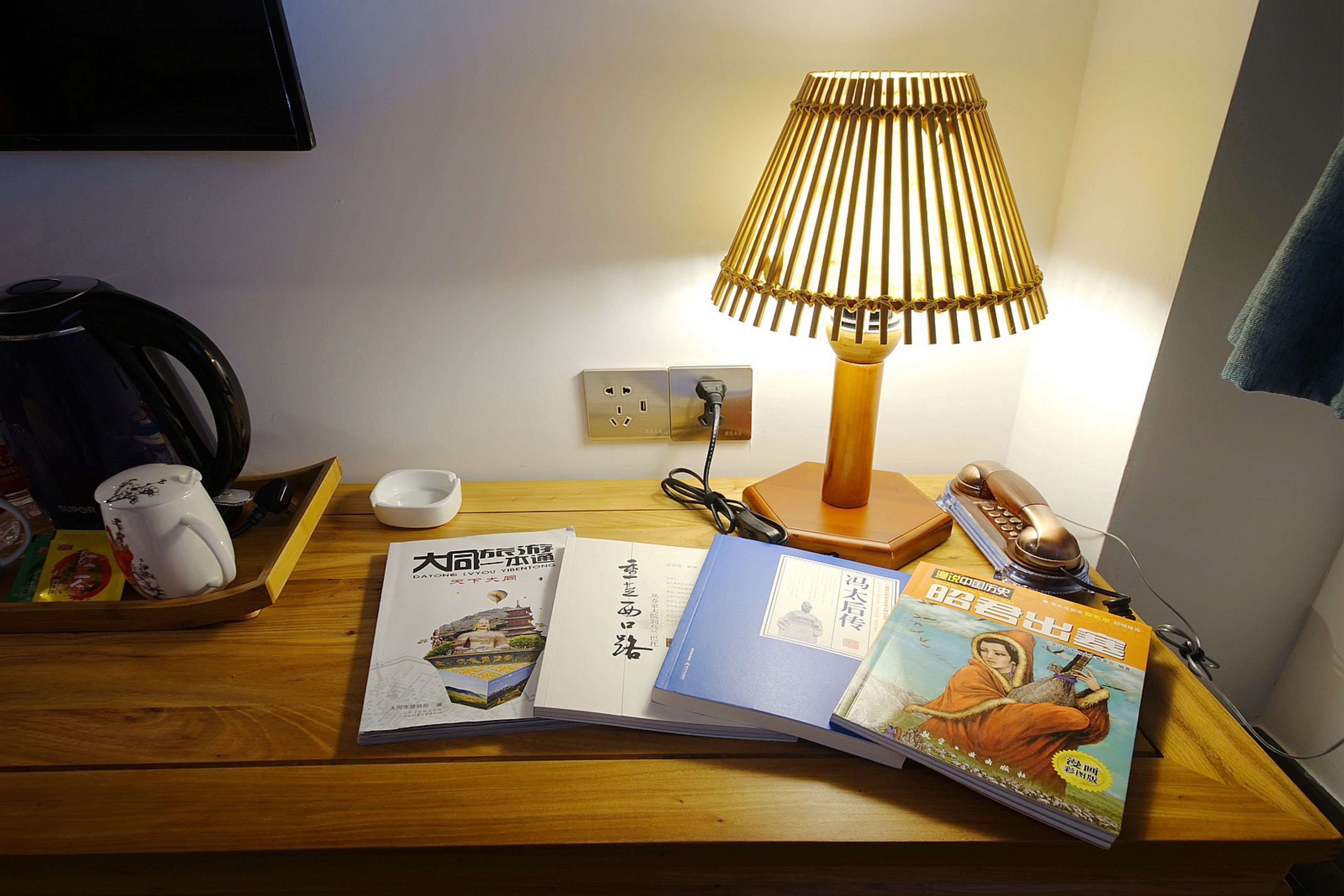 A wooden bedside table with a lamp, books, a mug, and a telephone.