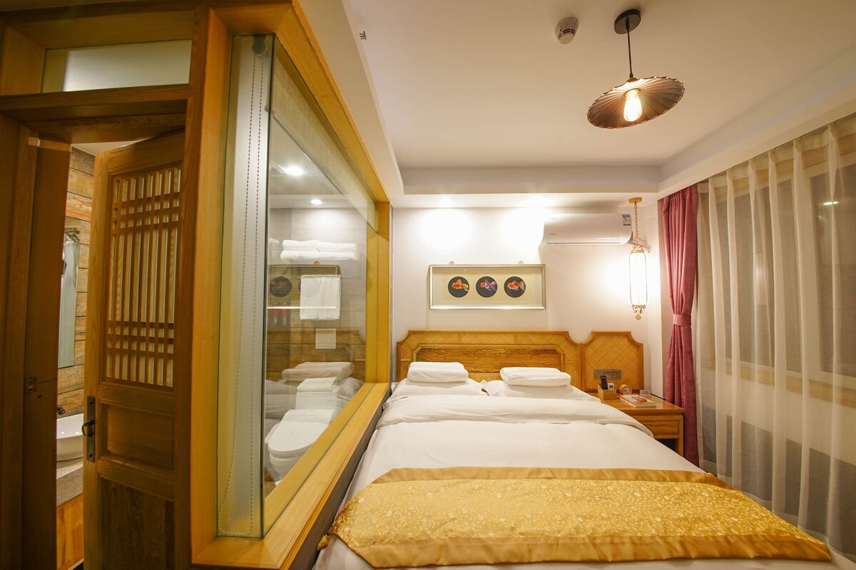 A hotel room with a large bed, a wooden headboard, a mirror, a bedside table with a lamp, and a window with curtains.
