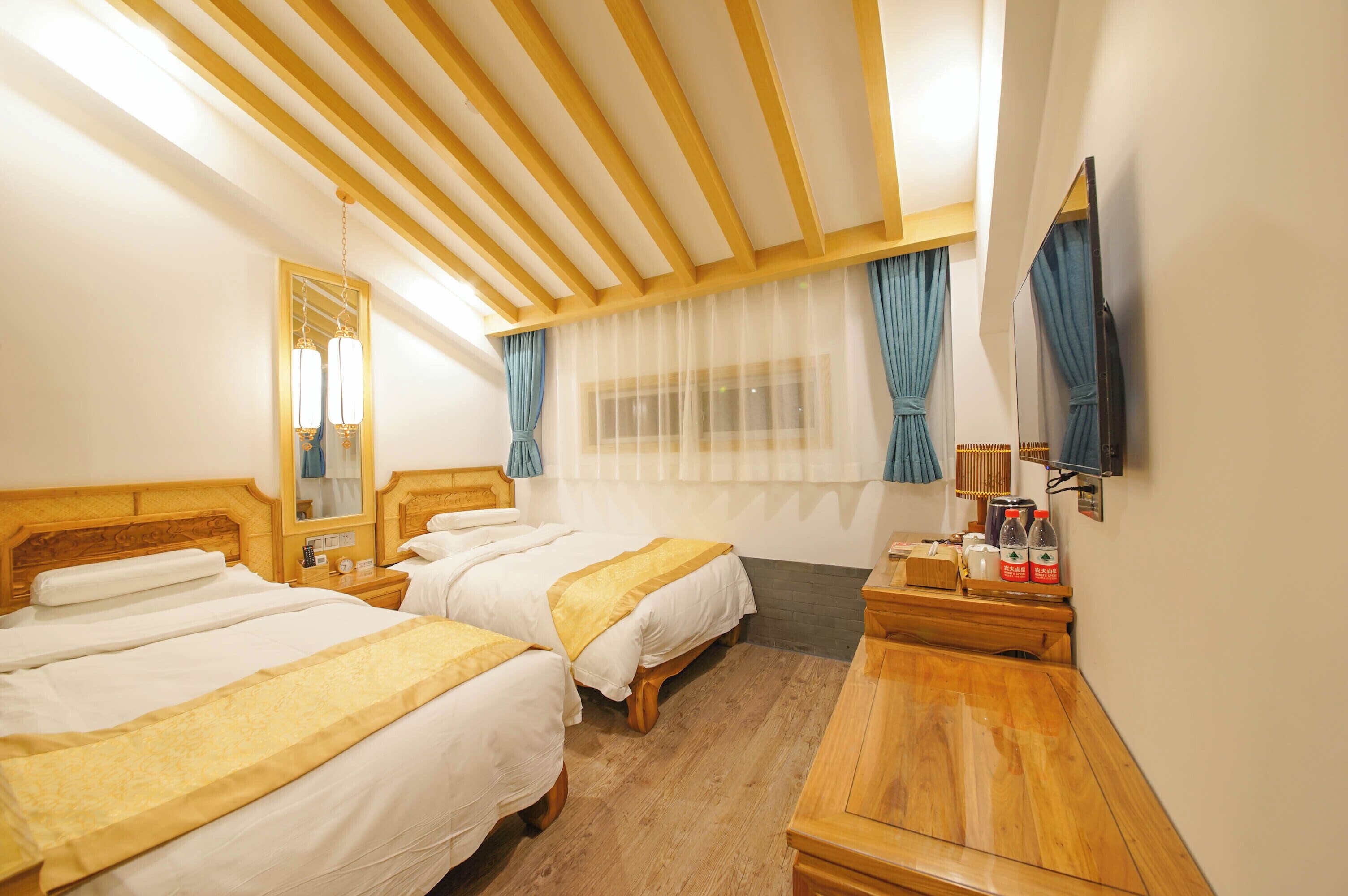 Habitación de hotel con dos camas, cabeceros de madera, una cómoda de madera, un espejo y una ventana con cortinas translúcidas.