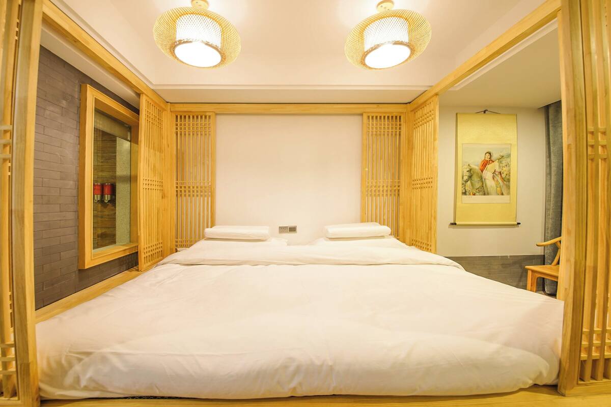 Una cama grande con sábanas blancas, cabecera de madera y mesitas de noche. Hay dos lámparas fijadas a la pared y un cuadro enmarcado.