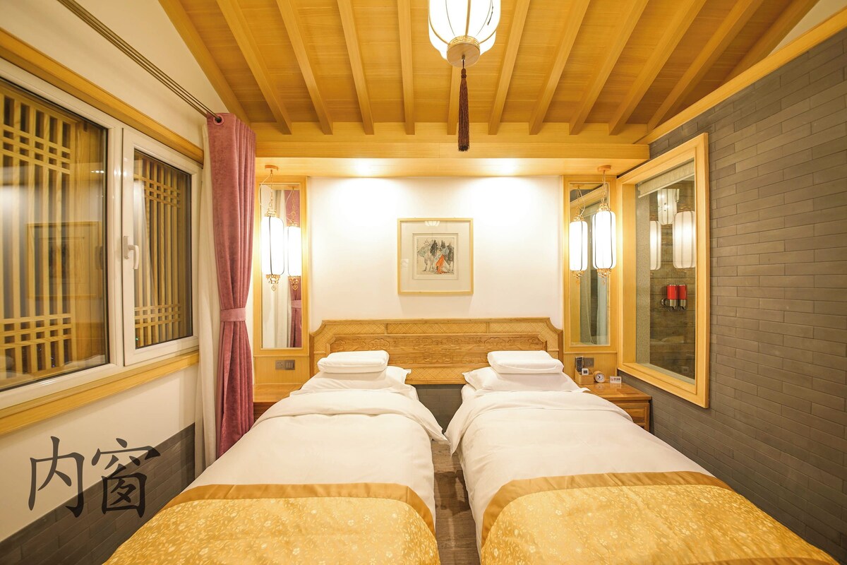 A hotel room with two beds, a wooden ceiling, a painting on the wall, and a window with a curtain.
