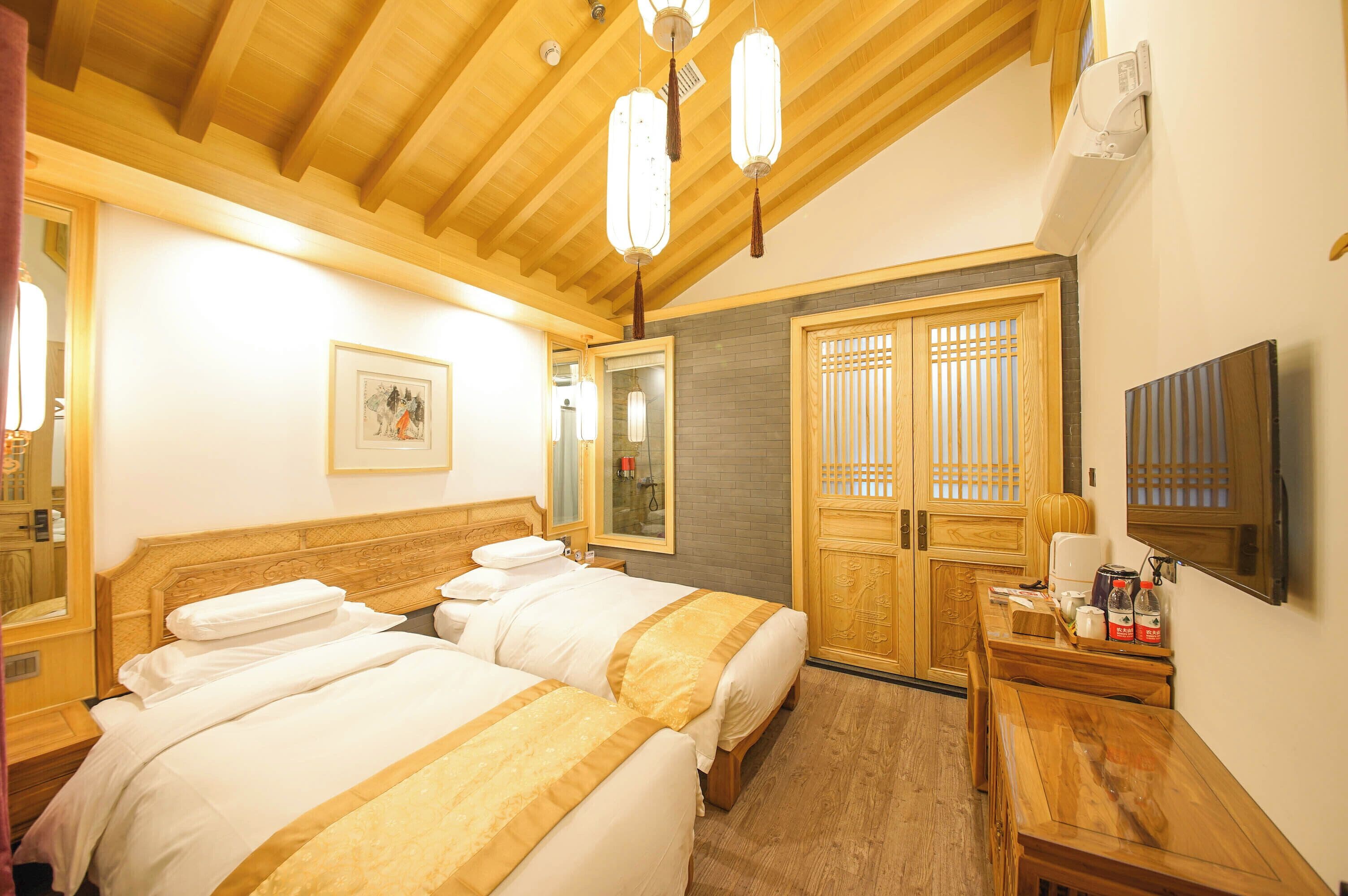 Een hotelkamer met een houten plafond, twee bedden met beddengoed in wit en geel, een houten bureau met een flatscreen televisie, en een traditionele houten deur met een ruit.