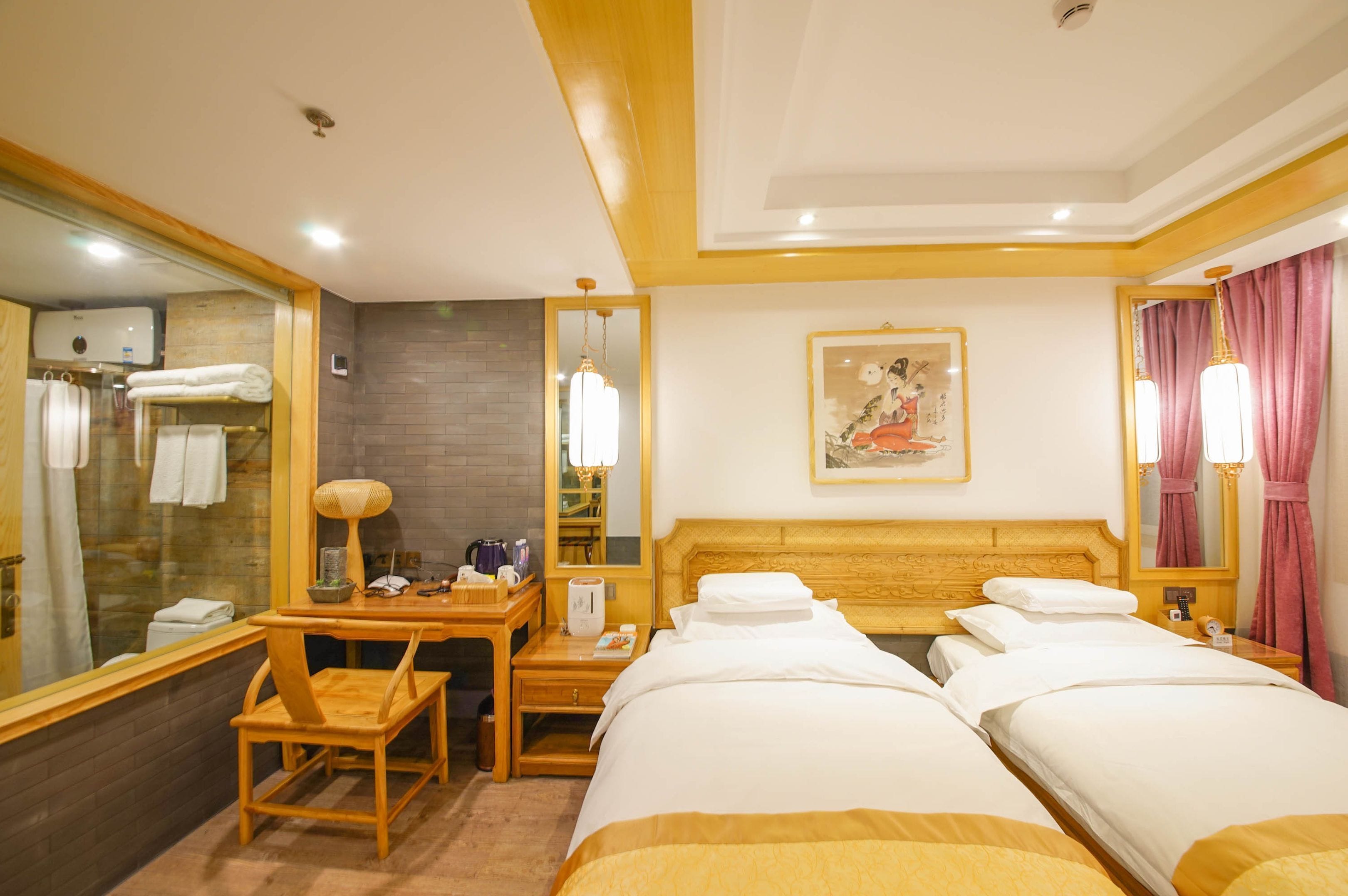 A hotel room with two beds, a desk with a chair, a mirror, a painting on the wall, and a bathroom visible through a glass partition.
