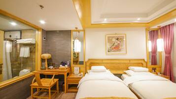 A hotel room with two beds, a desk with a chair, a mirror, a painting on the wall, and a bathroom visible through a glass partition.