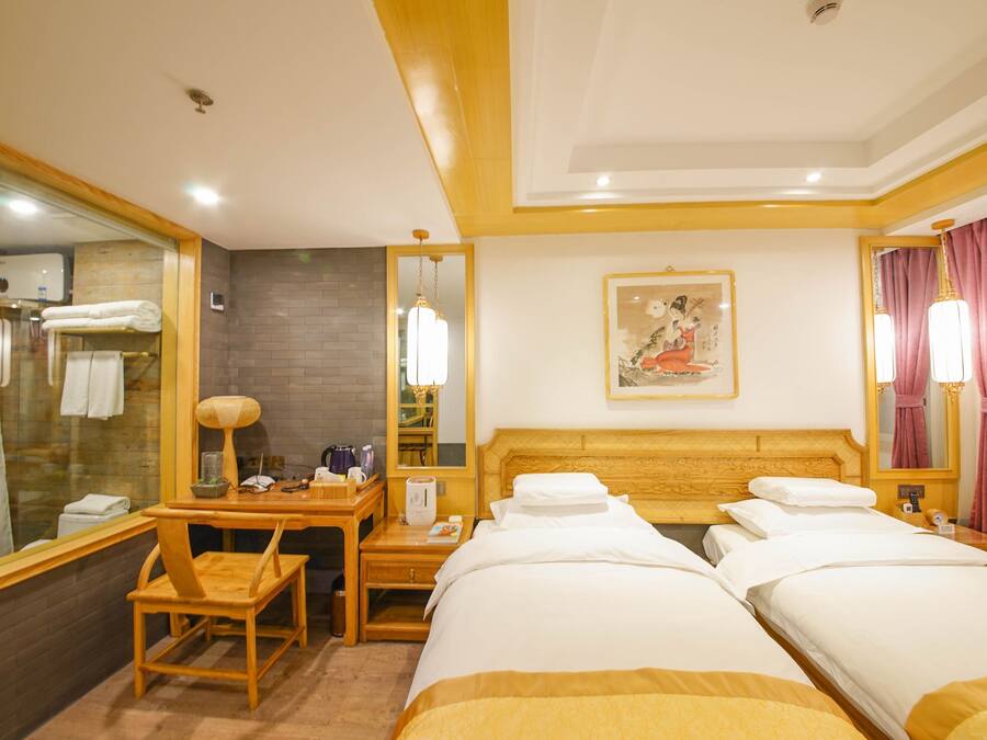 A hotel room with two beds, a desk with a chair, a mirror, a painting on the wall, and a bathroom visible through a glass partition.