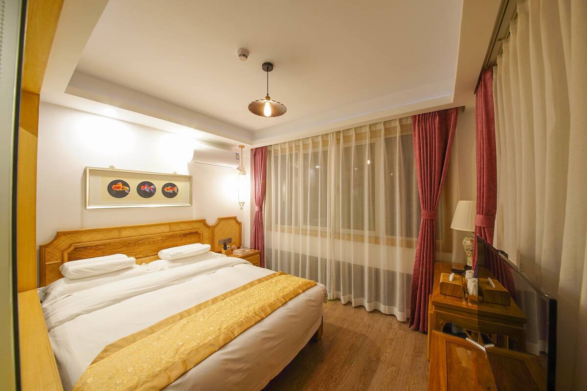 Habitación de hotel con una cama grande, una mesita de noche con lámpara, un espejo y una ventana con cortinas translúcidas.