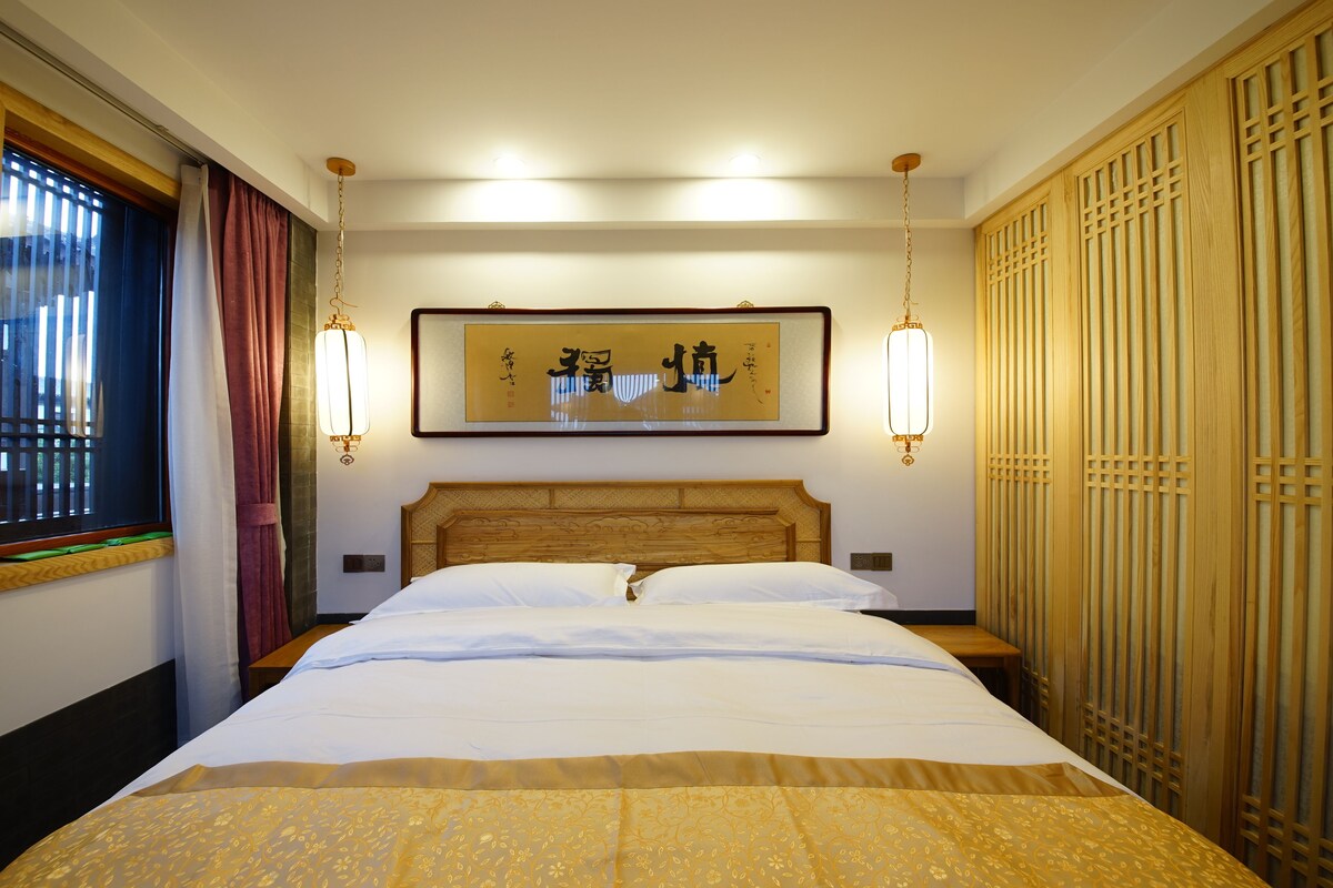 A hotel room with a large bed, wooden headboard, two wall-mounted lights, a framed artwork, and a wooden wardrobe.