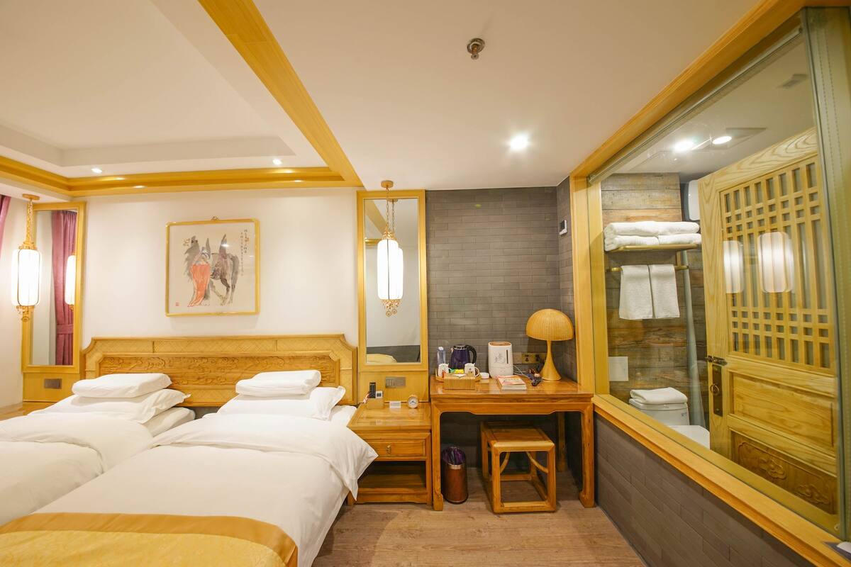 Habitación de hotel con dos camas, un escritorio, un espejo y un baño visible a través de un separador de vidrio.