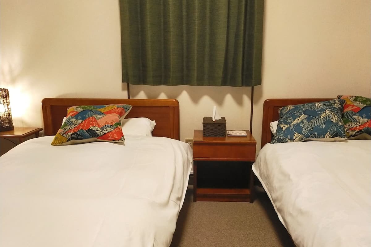 A hotel room with two beds, each with a wooden headboard, white bedding, and colorful pillows. There is a nightstand between the beds with a tissue box and a lamp. The room has a carpeted floor and a green curtain on the window.