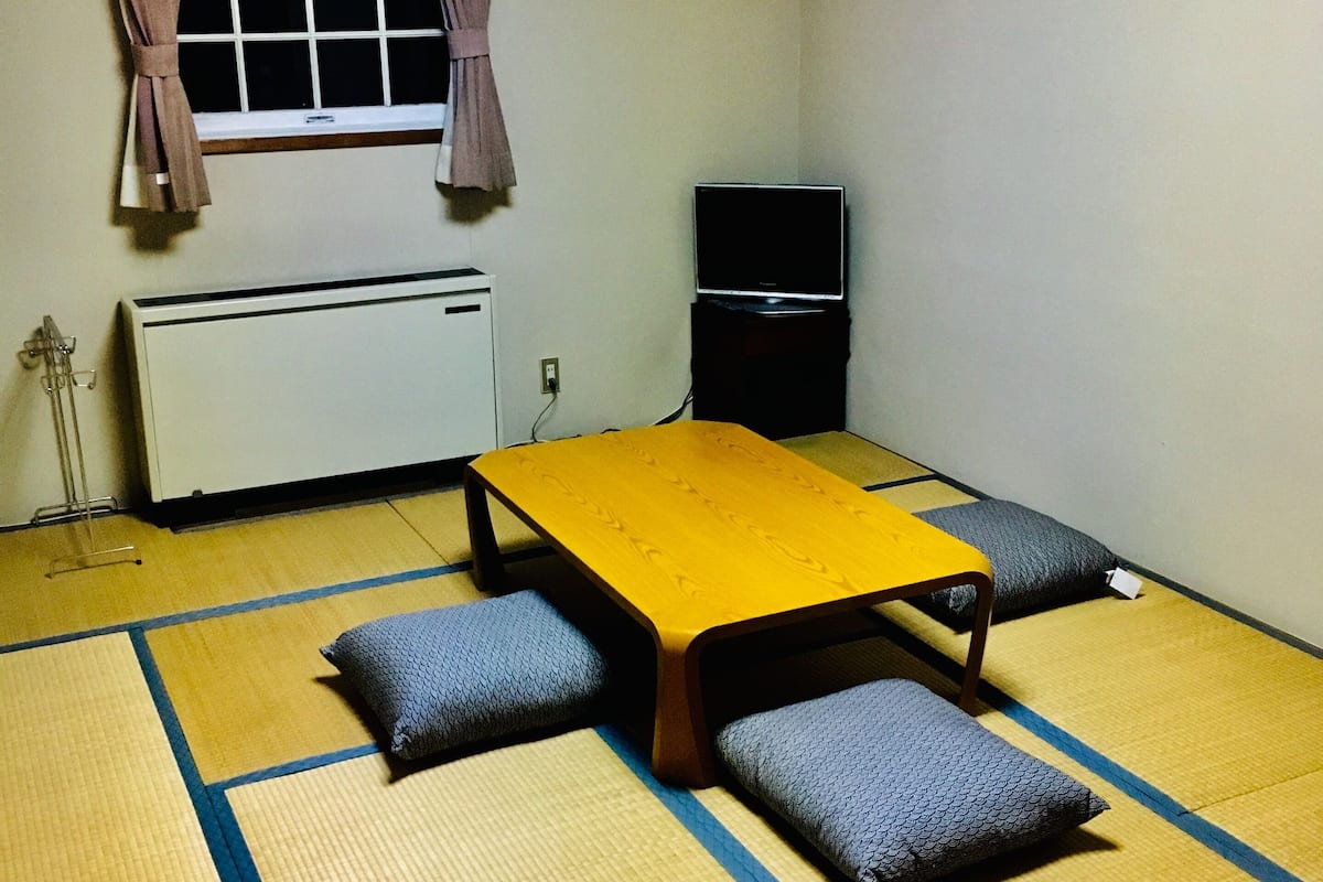 A room with tatami flooring, a low yellow table, two cushions, a black speaker, and a window with curtains.