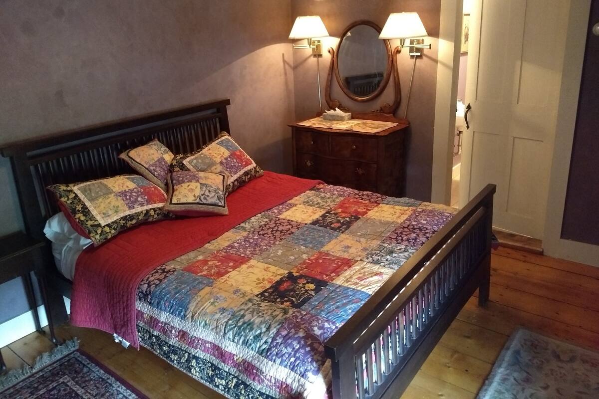 A bedroom with a wooden bed, a red blanket, a floral quilt, and two wall-mounted lamps.