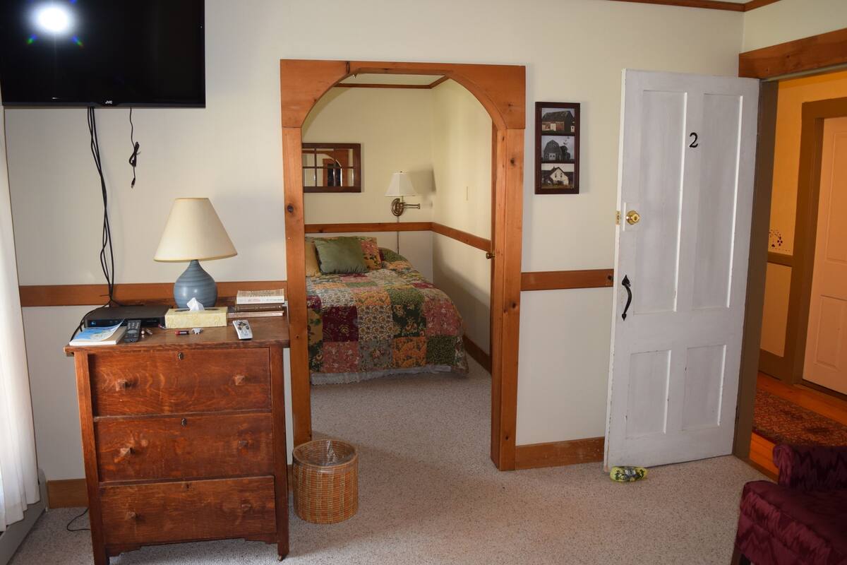 A bedroom with a wooden dresser, a lamp, a bed with a quilt, a television mounted on the wall, and a door with the number 2.
