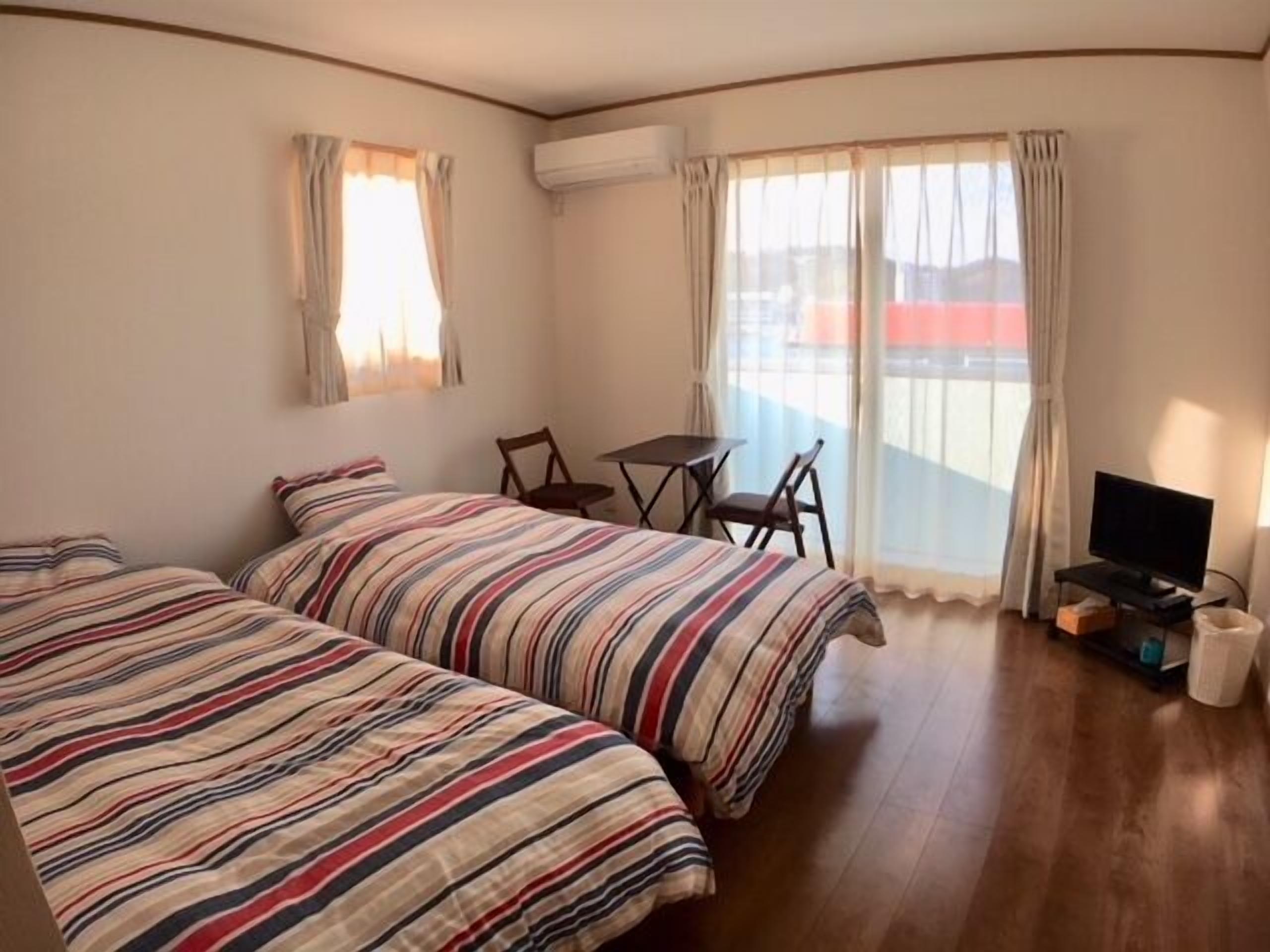 A hotel room with two beds, a small table, chairs, a television, and a balcony with curtains.