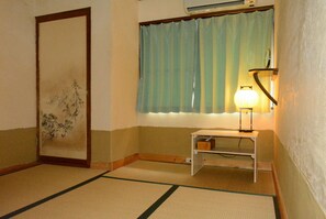 A room with tatami flooring, a shoji screen door, a window with blue curtains, and a bedside table with a lamp. - Guesthouse Bon (Kyoto)